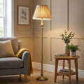 Flemish brass floor lamp with a pleated beige shade standing next to a grey sofa in a warm living room setting.