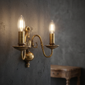 Two-light antique brass wall sconce with glowing candle bulbs mounted on a textured grey wall.