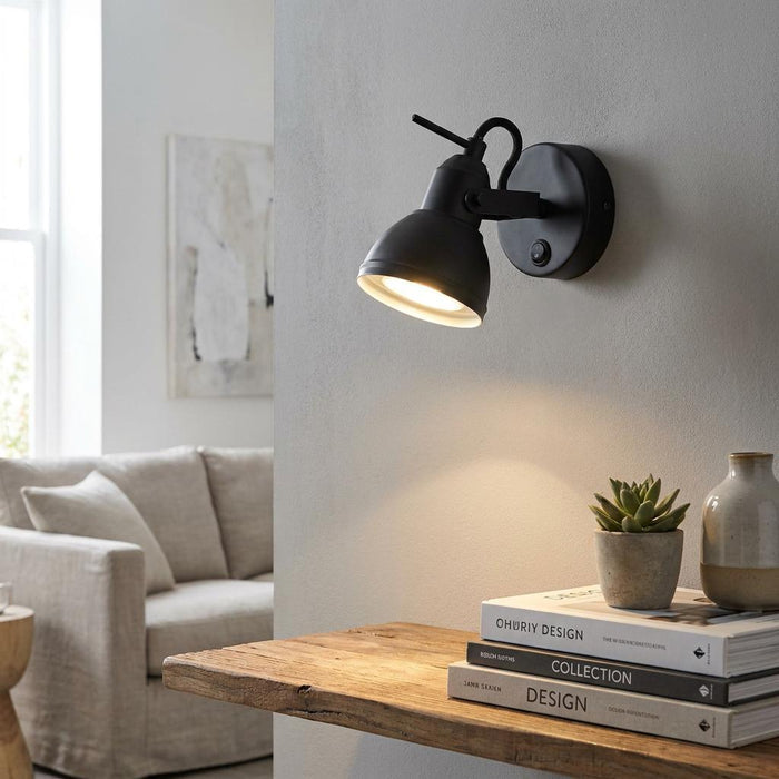 Black adjustable wall spotlight mounted on a grey wall, illuminating a shelf with books and a small plant.
