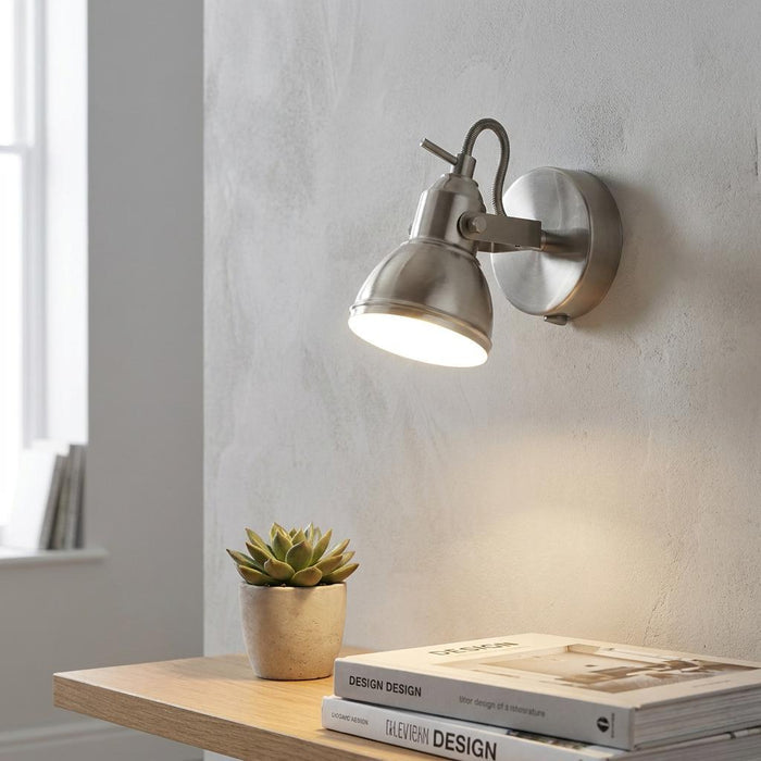 Brushed silver adjustable wall spotlight illuminating a potted succulent and books on a wooden shelf.