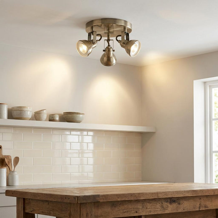 Three-lamp antique brass spotlight fixture mounted on a kitchen ceiling above a wooden island and white tiled wall.