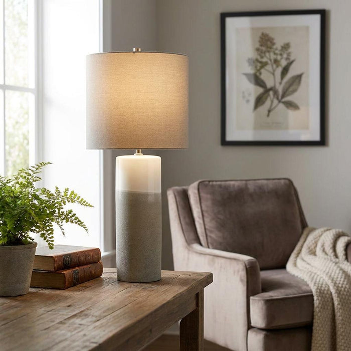 Two-tone ceramic table lamp with a grey drum shade on a rustic wooden table in a living room setting.