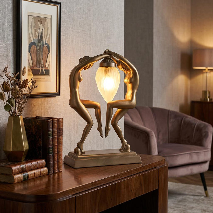 Art Deco table lamp with two golden female figures holding a glowing frosted glass teardrop shade on a wooden table.