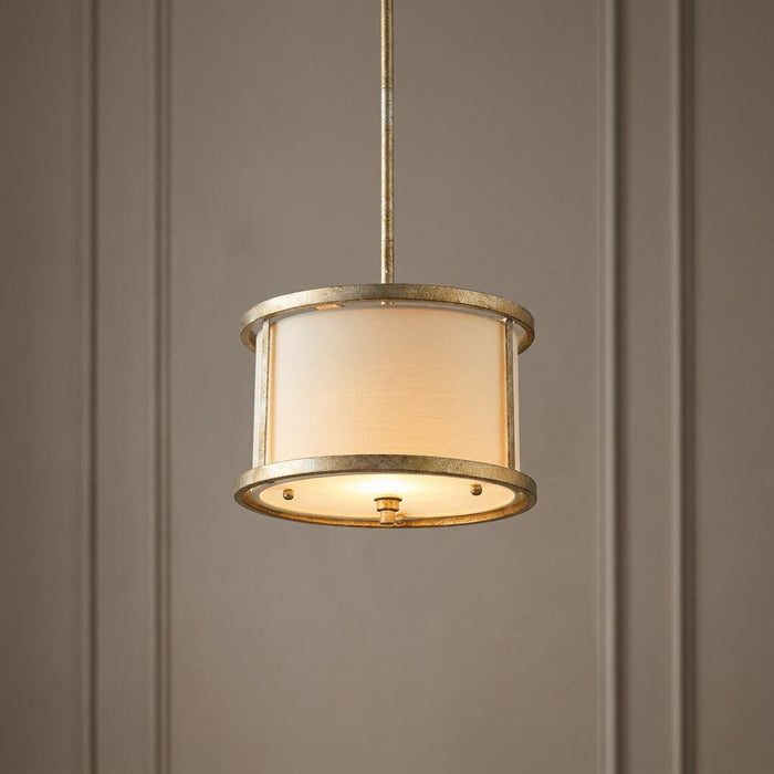 Gilded mini pendant light with a cream drum shade and textured metallic frame against a paneled wall.