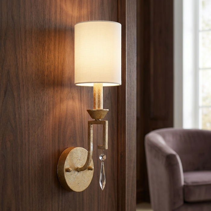 Gilded Nola Lemuria gold wall light with a white fabric shade and crystal drop on a wood panel wall.