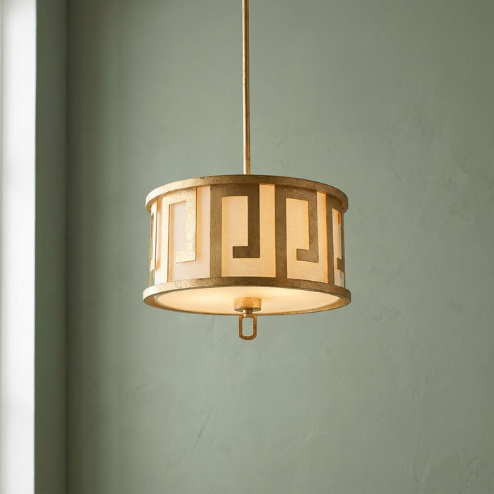 A gilded drum pendant with a Greek key pattern and fabric shade hanging against a muted green wall.