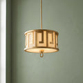 A gilded drum pendant with a Greek key pattern and fabric shade hanging against a muted green wall.