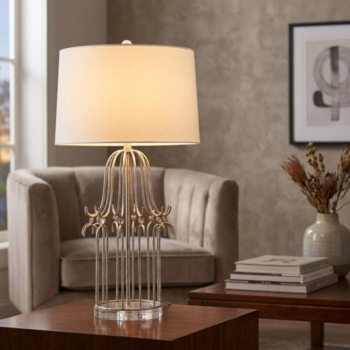Distressed silver table lamp with a cage-style base and white drum shade on a wooden table in a living room.