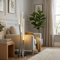 A three-light chrome floor lamp with glowing bubble glass tubes stands next to a grey armchair in a modern living room.