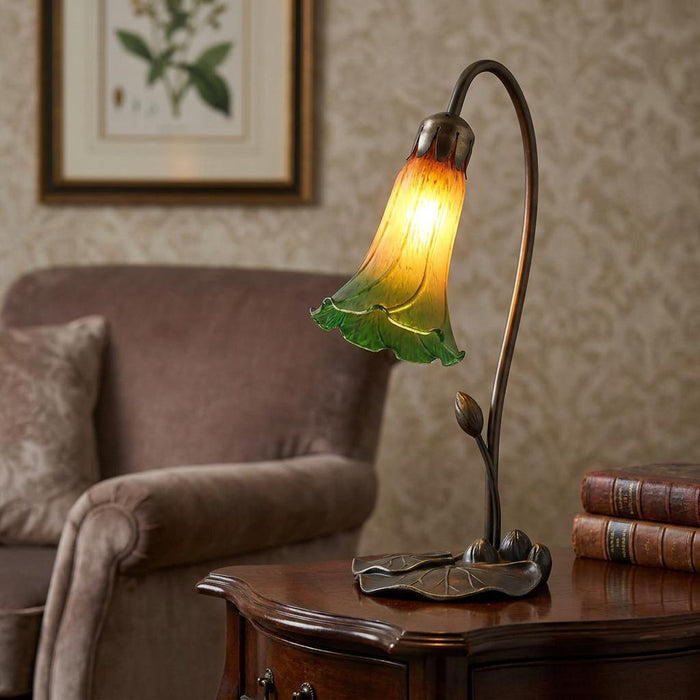 Green and amber lily glass table lamp with a bronze stem base glowing on a wooden table in a cozy living room.