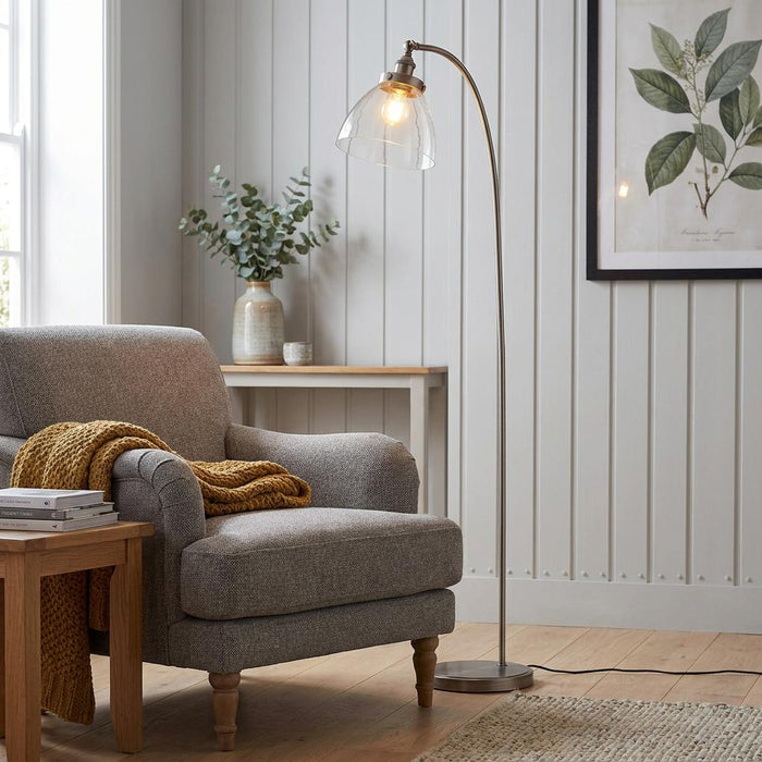 Brushed steel floor lamp with a clear glass shade positioned next to a grey armchair in a cozy living room.