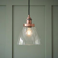 Copper pendant light with a clear glass dome shade and glowing bulb against a sage green paneled wall.