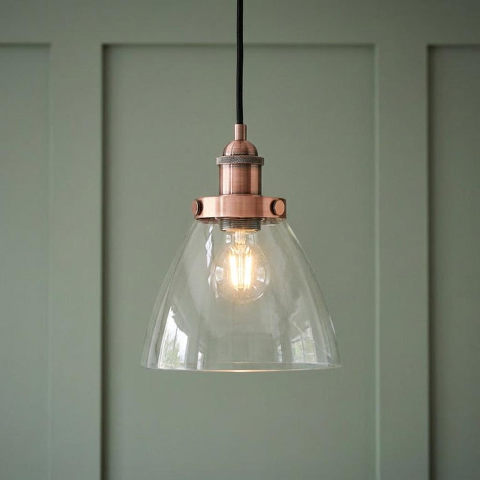 Copper pendant light with a clear glass dome shade and glowing bulb against a sage green paneled wall.