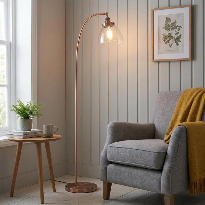 Arched copper floor lamp with a glass shade illuminating a cozy living room nook with a gray armchair.