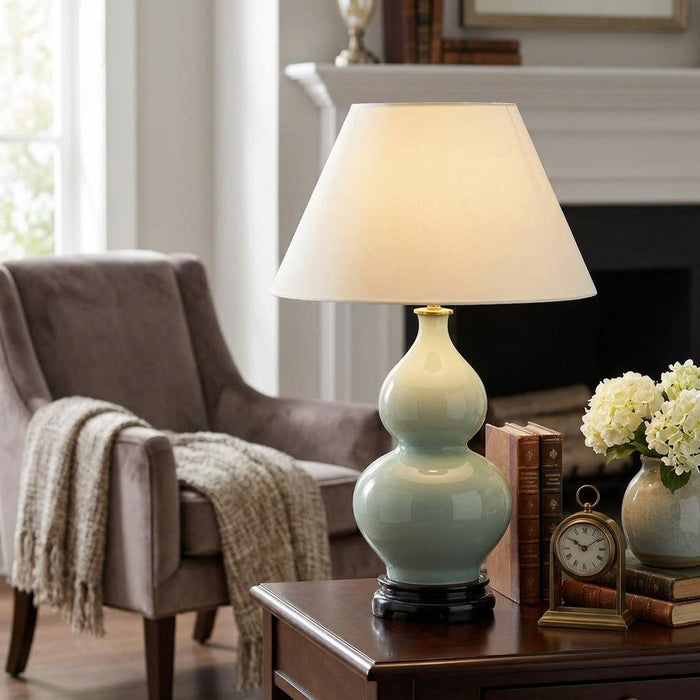 Lit celadon ceramic double-gourd table lamp with an off-white shade on a dark wooden side table.