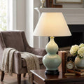 Lit celadon ceramic double-gourd table lamp with an off-white shade on a dark wooden side table.