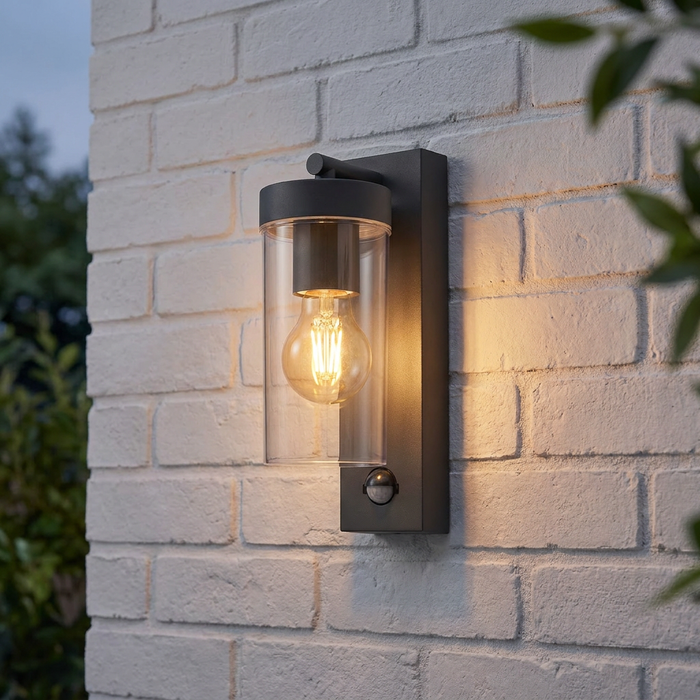Hayden PIR sensor outdoor wall light with a warm lit bulb in a cylindrical glass shade on a white brick wall.