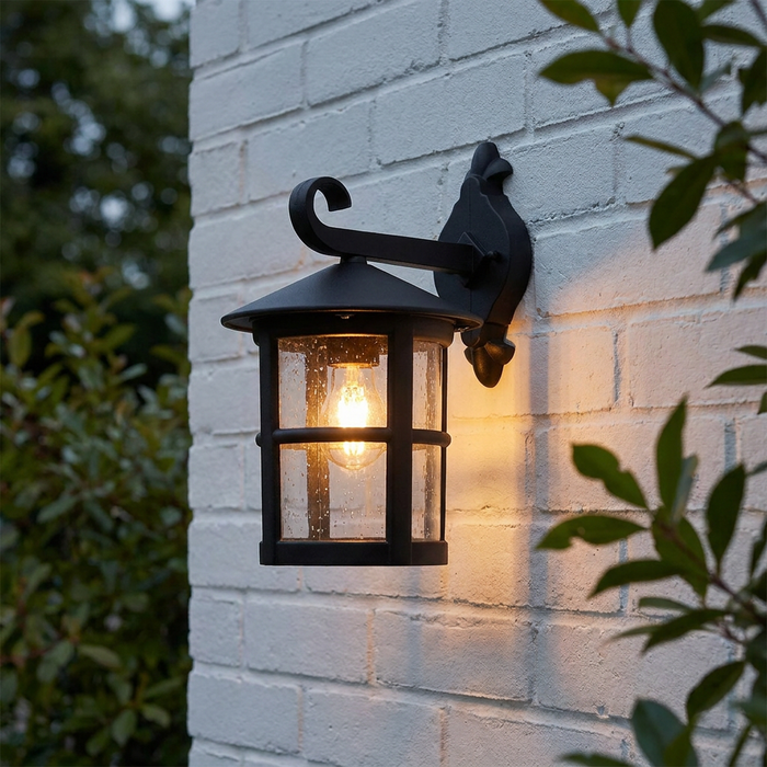 Hereford II black outdoor wall lantern with seeded glass and a warm bulb mounted on a white brick wall.