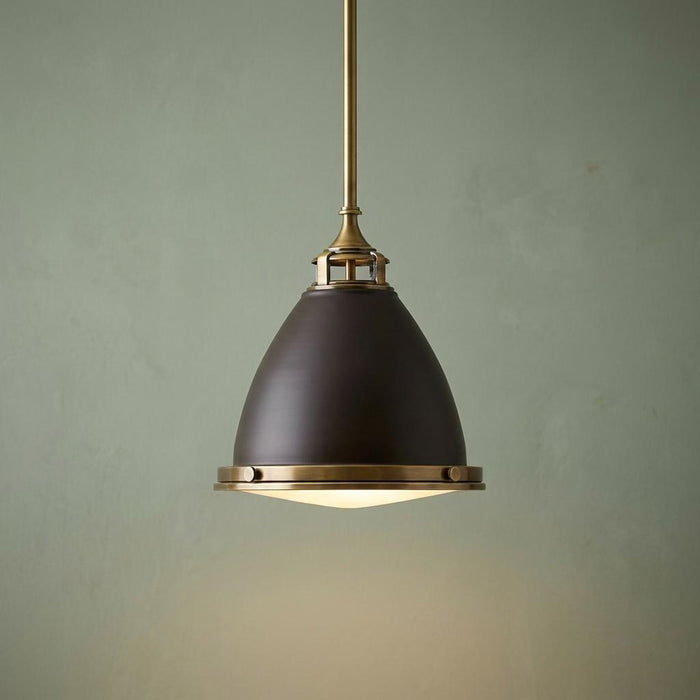 Industrial-style bronze and brass pendant light with a bell-shaped shade hanging against a muted green wall.