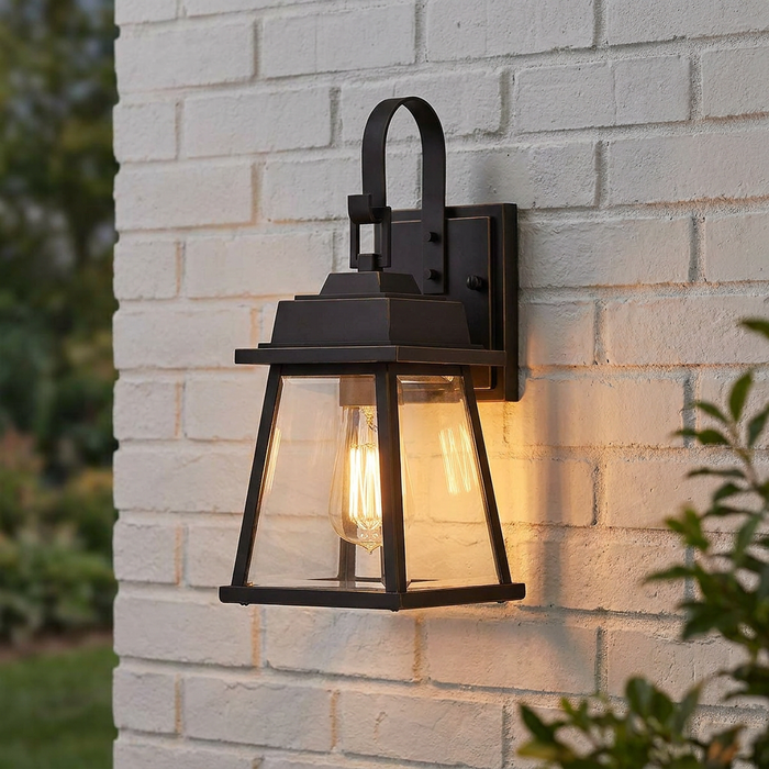 Bronze outdoor wall lantern with a glowing bulb mounted on a white painted brick wall.