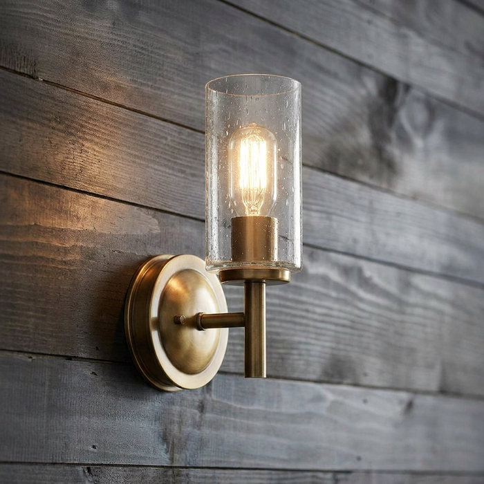Heritage brass wall light with a cylindrical seeded glass shade and Edison bulb on a rustic wood plank wall.