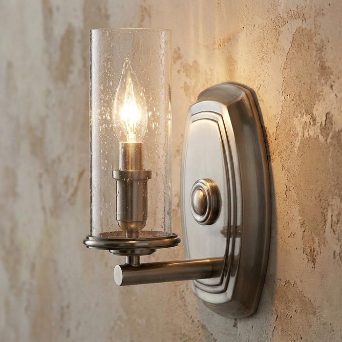 Polished antique nickel wall sconce with a cylindrical seeded glass shade and a warm candle bulb.