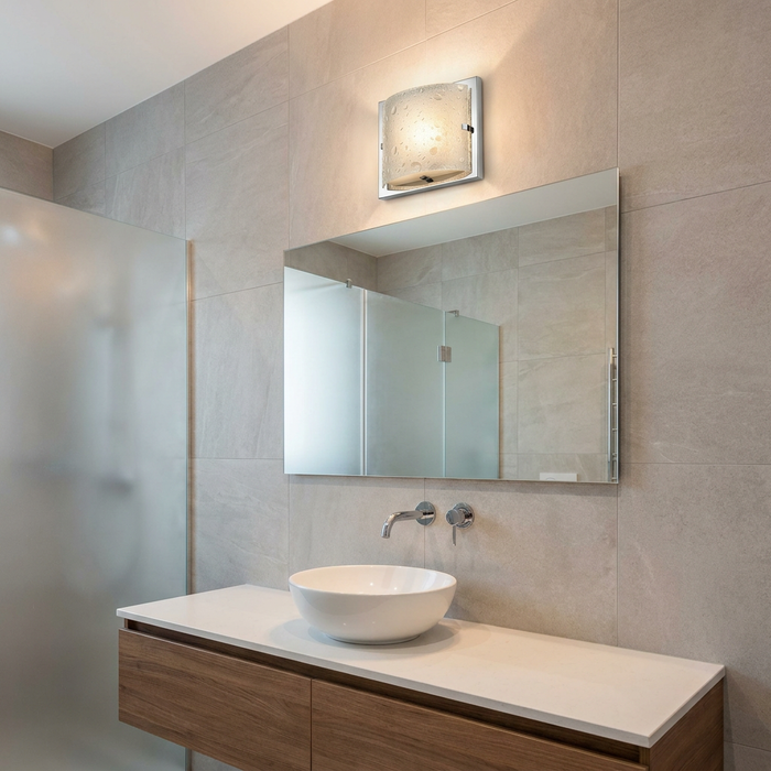 Polished chrome bathroom wall light with seeded glass shade above a mirror in a modern tiled bathroom.