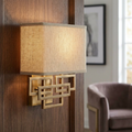 Brushed bronze wall light with geometric interlocking rectangles and a linen shade on a wood-paneled wall.