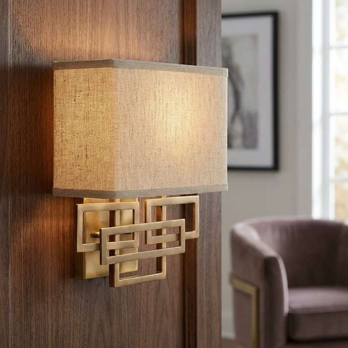 Brushed bronze wall light with geometric interlocking rectangles and a linen shade on a wood-paneled wall.