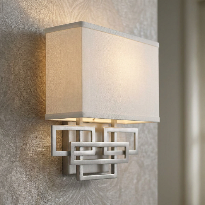 Wall light with a white rectangular fabric shade and an interlocking geometric nickel base on a patterned wall.
