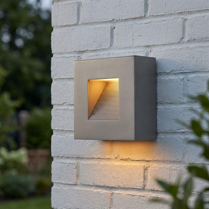 Modern square titanium outdoor wall light with a warm glow mounted on a white brick exterior wall.