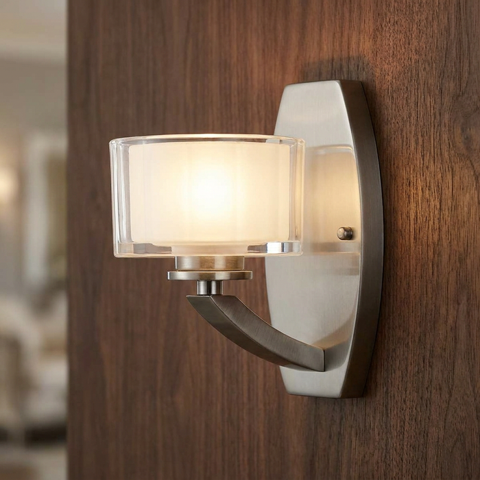 Brushed nickel wall light with a dual glass shade mounted on a dark wood-paneled wall.