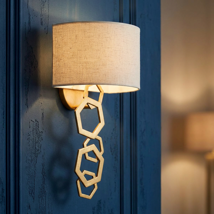 Hinkley Moxie gold wall light with linen shade and geometric hexagon detail on a navy blue paneled wall.
