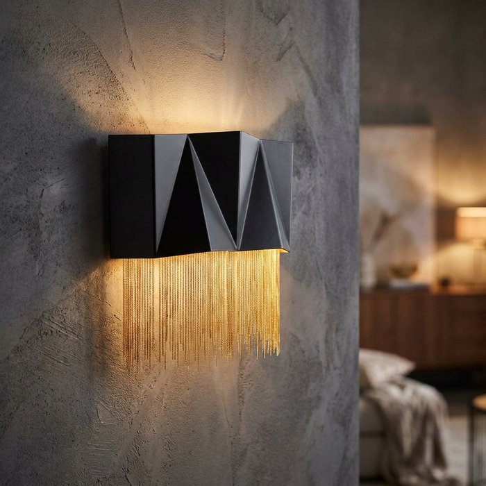 Modern black geometric wall light with gold chain fringe mounted on a textured grey wall, emitting warm light.