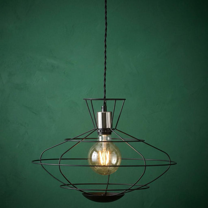Industrial black cage pendant light with a chrome bulb holder and glowing Edison bulb against a dark green wall.