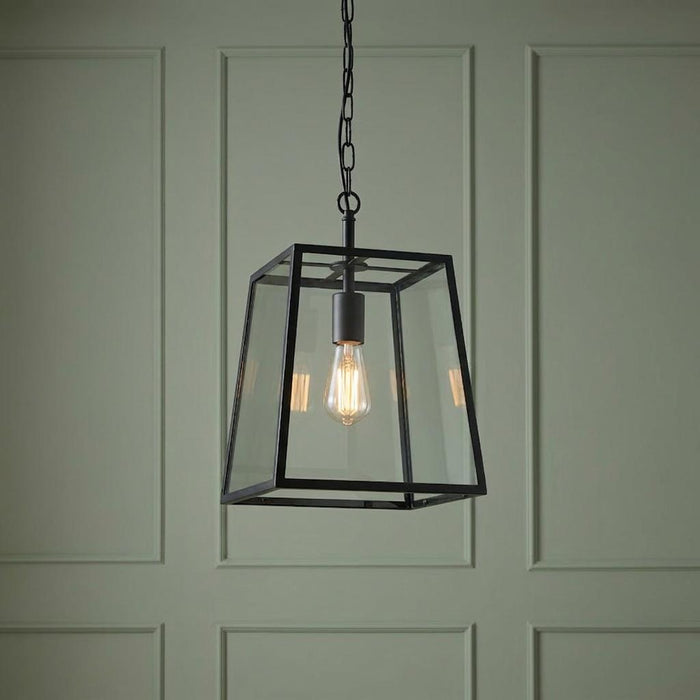 Black lantern-style pendant light with a clear glass shade and exposed bulb against a paneled sage green wall.