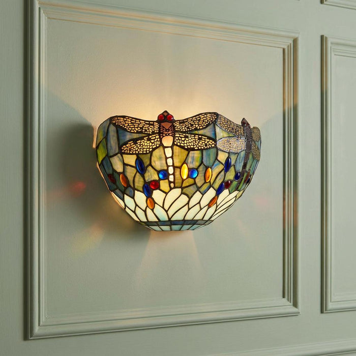 Blue dragonfly Tiffany-style stained glass wall light illuminated on a pale green panelled wall.