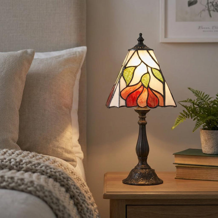 A Tiffany bedside lamp with a floral stained glass shade glowing on a wooden nightstand next to a bed.
