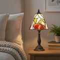 A Tiffany bedside lamp with a floral stained glass shade glowing on a wooden nightstand next to a bed.