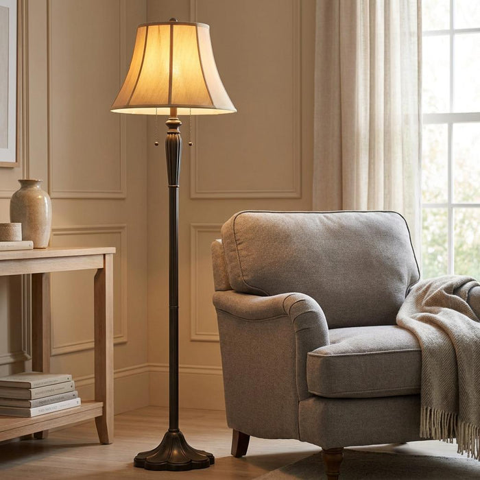 A dark bronze floor lamp with a pleated beige shade stands beside a grey armchair in a cozy living room.