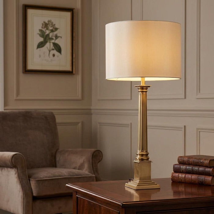 Solid brass fluted column table lamp with a cream drum shade sitting on a wooden desk beside leather books.