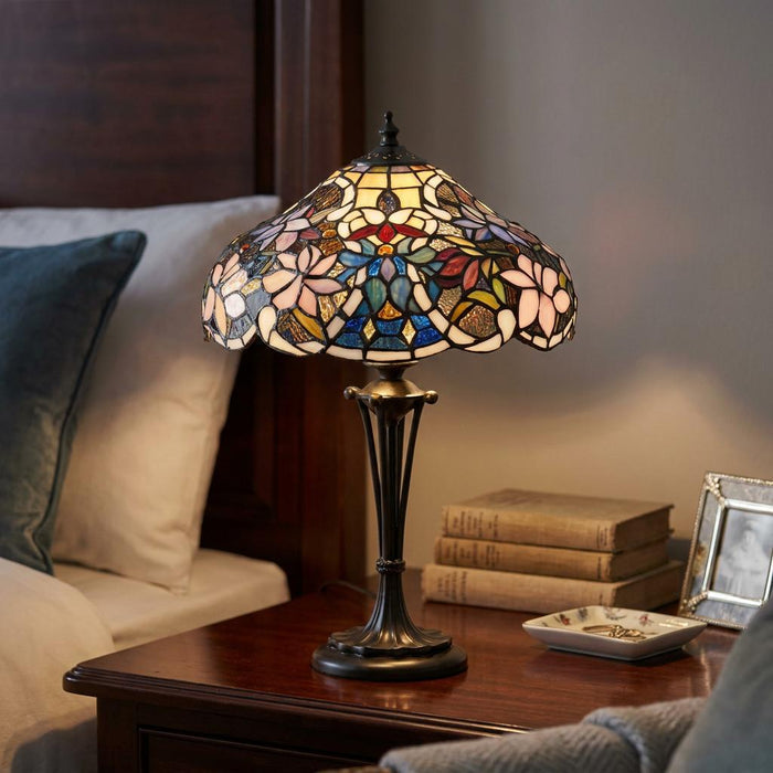 Ornate floral Tiffany bedside lamp glowing on a dark wood nightstand next to pillows and antique books.