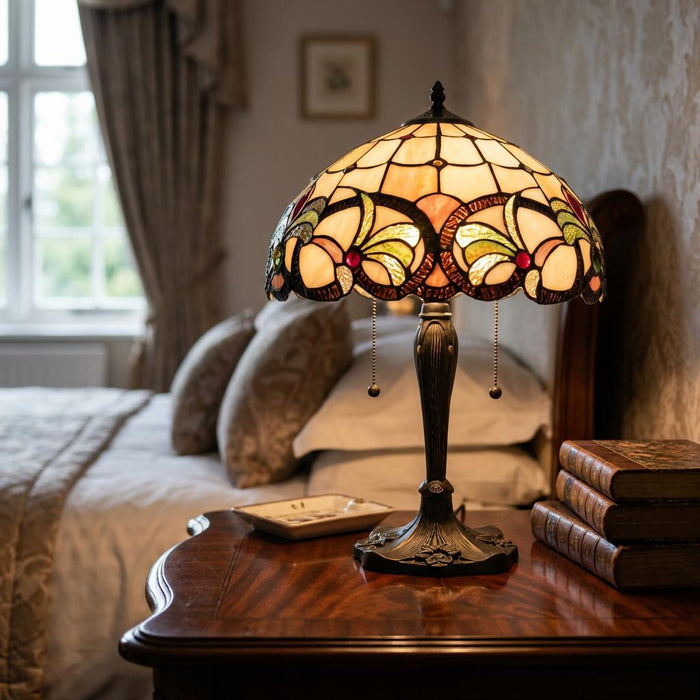 Ipswich Tiffany table lamp with a floral stained glass shade illuminated on a wooden bedside table in a bedroom.