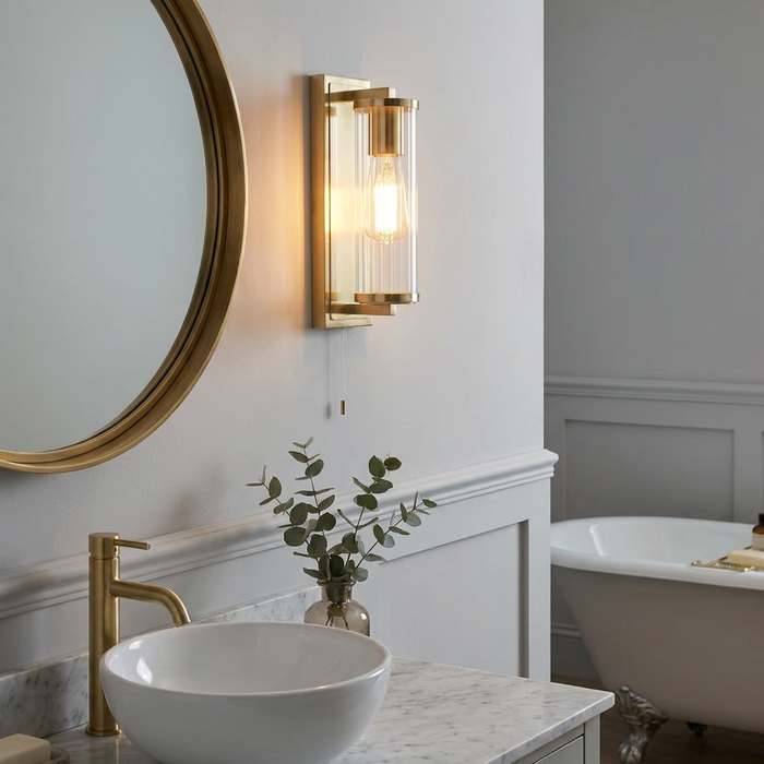 Brass Art Deco wall light with a fluted glass shade mounted above a marble bathroom vanity next to a round mirror.