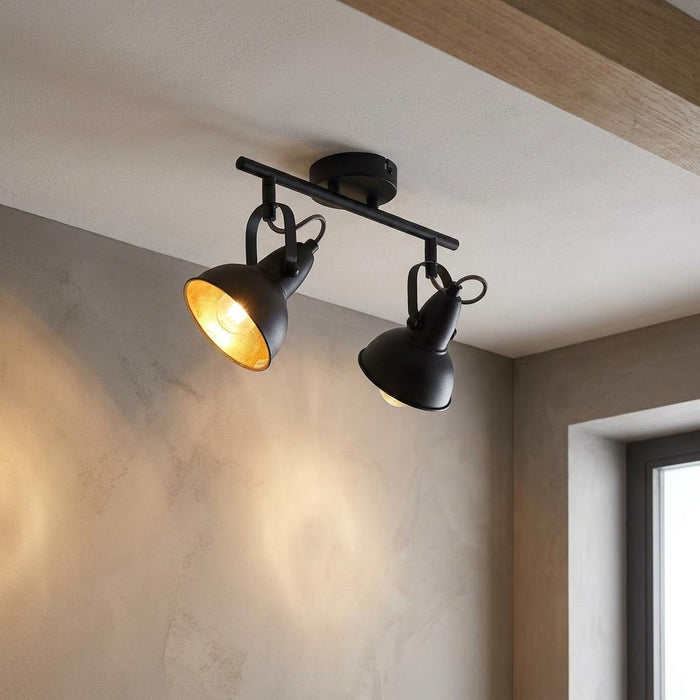 Jurva Urban black double bar spotlight with gold inner shades, mounted on a ceiling and illuminating a textured wall.