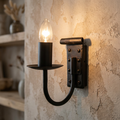 Rustic black wrought iron candle-style wall sconce with a lit bulb against a textured plaster wall.