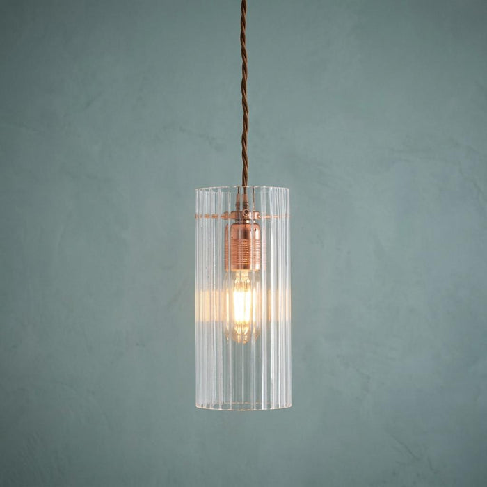 Lit Kansa elongated prismatic ceiling light with copper bulb holder and clear ribbed shade on a textured teal wall.