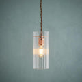Lit Kansa elongated prismatic ceiling light with copper bulb holder and clear ribbed shade on a textured teal wall.
