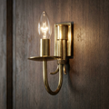 A polished brass single wall light with a glowing candle bulb mounted on a dark wood panel.
