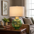 Kara table lamp with a green glass globe base and white drum shade on a wooden side table in a cozy living room.